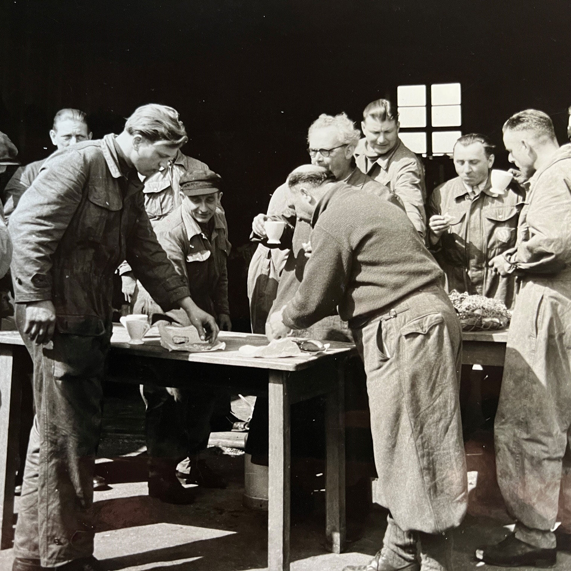 Black and white picture of a few people standing around a table looking at things lying on the table.