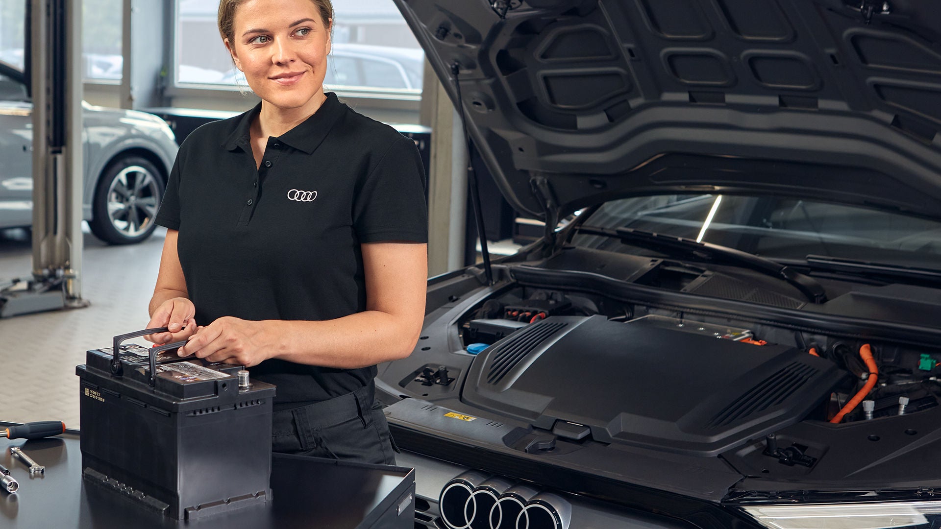 Μια γυναίκα στέκεται σε ένα συνεργείο μπροστά από ένα Audi με το καπό ανοιχτό, κρατώντας μια μπαταρία εκκίνησης.