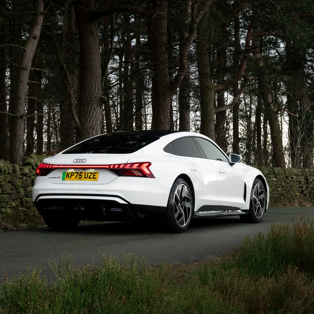 White Audi e-tron GT parked on a quiet country road, shown from the rear three-quarter angle. The vehicle features a sleek fastback silhouette, large alloy wheels and a continuous rear LED light bar. A stone wall and tall pine trees line the roadside, creating a natural woodland backdrop under overcast daylight.