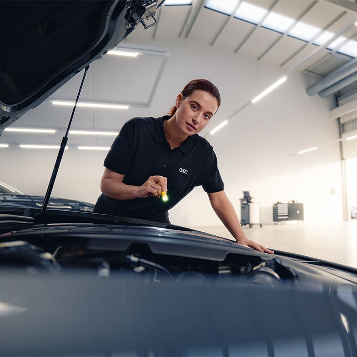 Une personne souriante, portant un polo noir, travaille sous le capot d’une voiture dans un atelier lumineux, donnant l’impression d’inspecter ou de réparer le moteur.