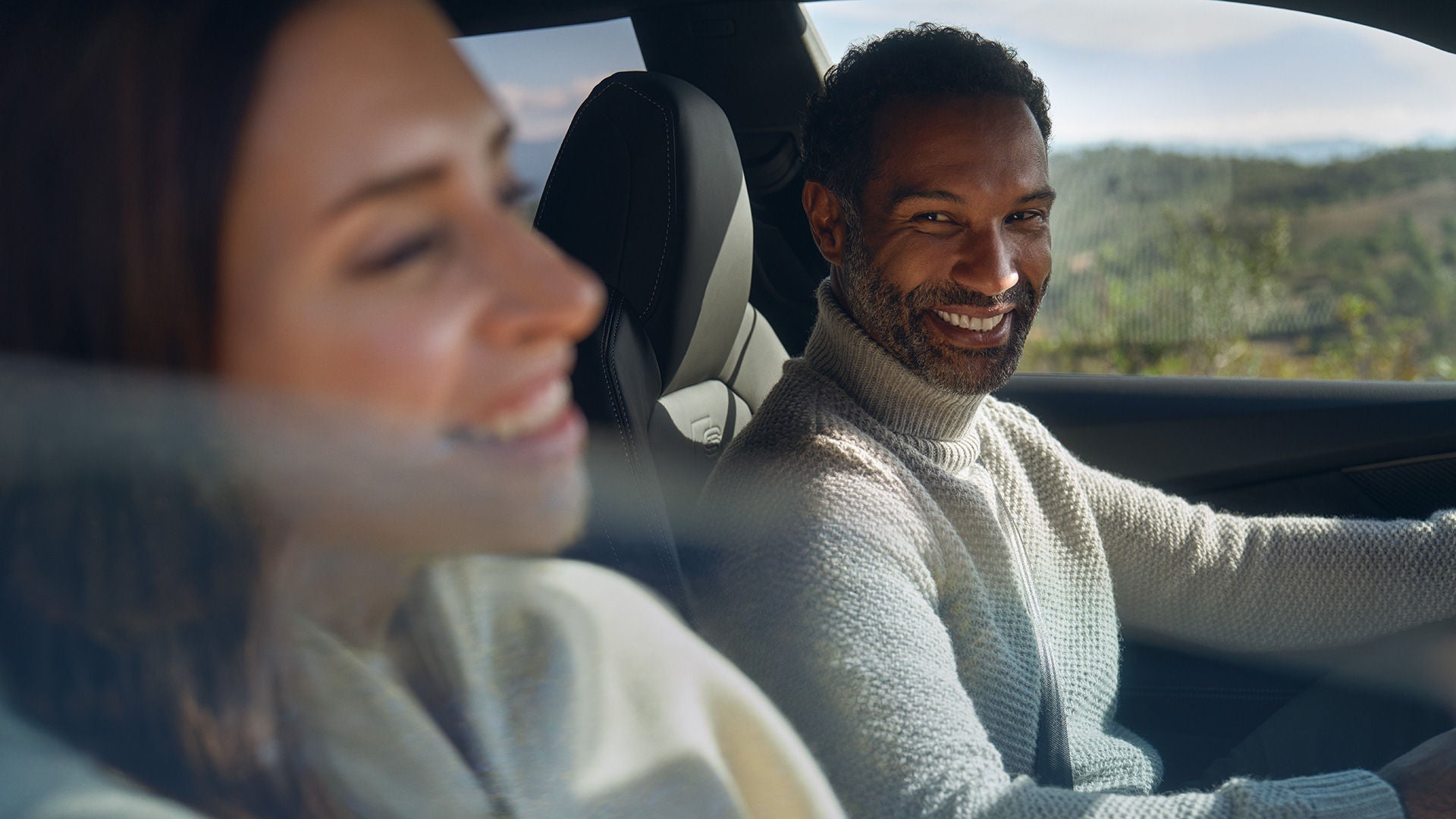 Mann und Frau sitzen lachend im Auto, der Mann schaut zur Kamera, durch das Fenster ist eine hügelige Landschaft zu sehen.