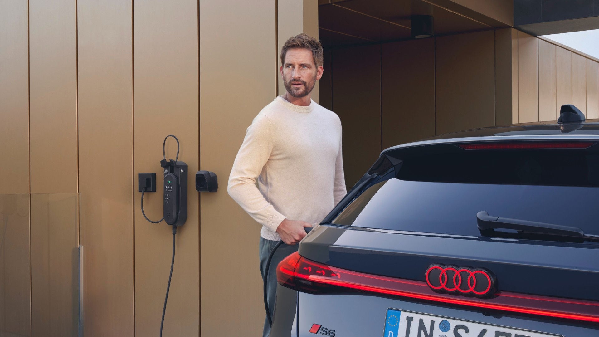 Man in a white sweater stands near a black Audi parked beside a brown wall with an electric charger.