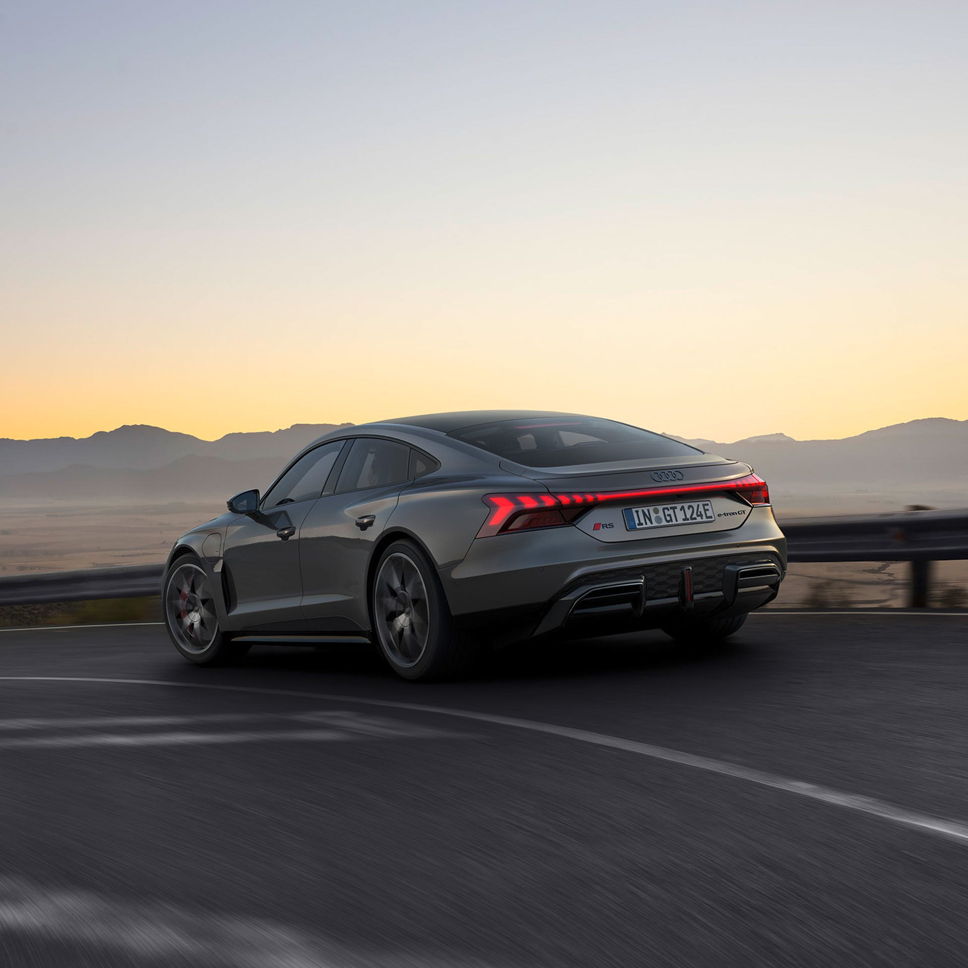 Ein grauer Audi RS e-tron GT fährt auf einer kurvigen Bergstraße bei Sonnenuntergang, mit leuchtenden Rücklichtern.