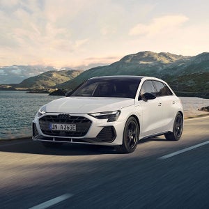 A white Audi drives along a scenic road beside a tranquil lake and mountains under a partly cloudy sky, evoking a sense of adventure and freedom.