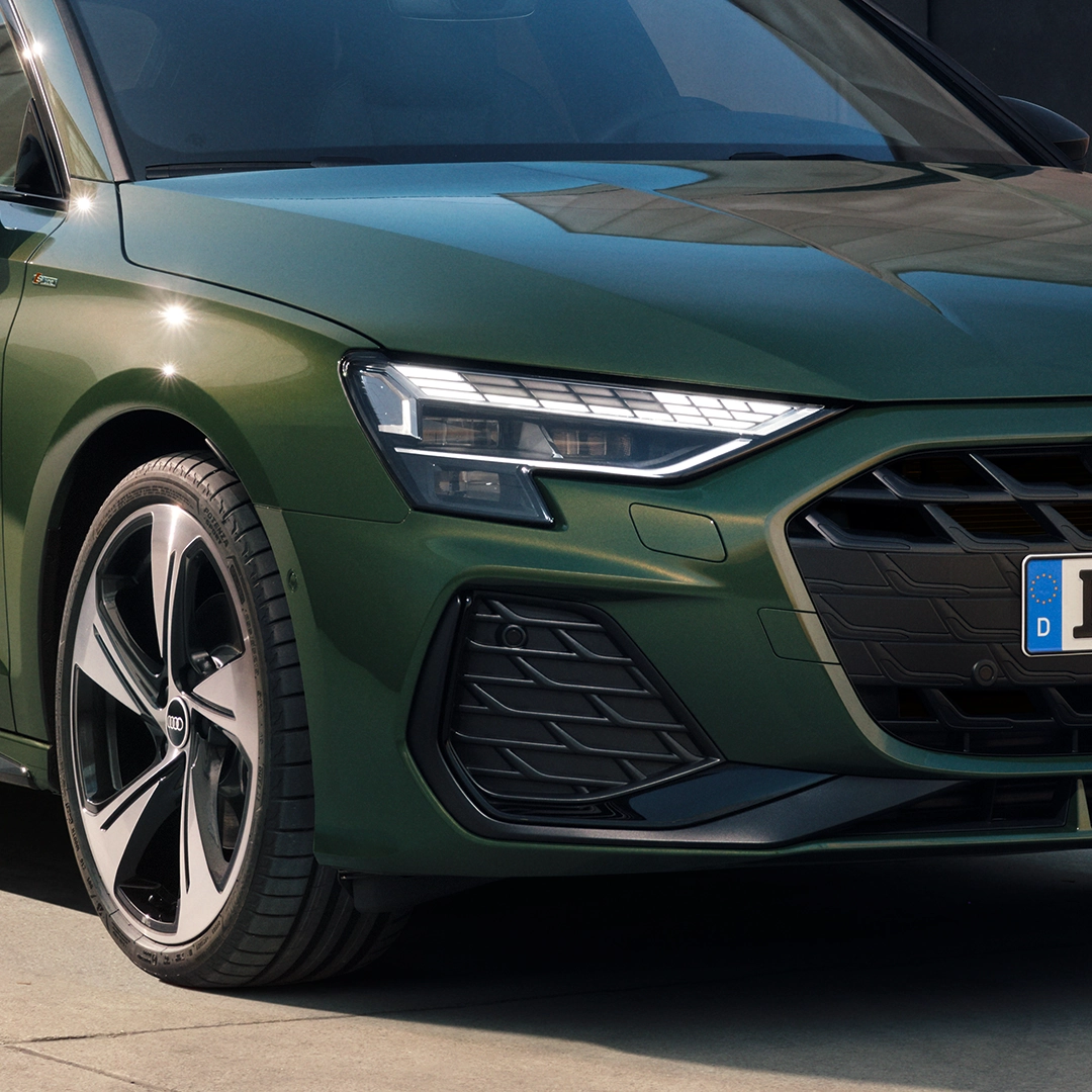 A portrait image of a green Audi A3 Sportback, close-up of front, showing headlight, grill, wheel, and part of windscreen.