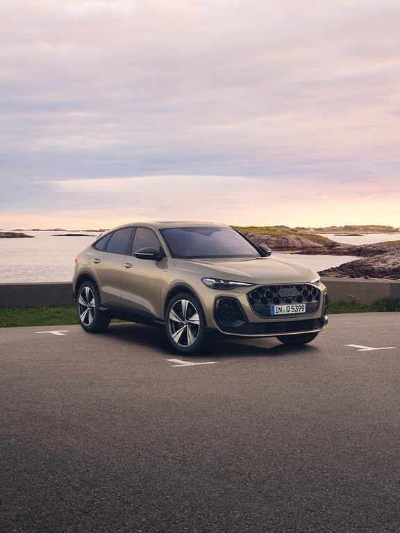 Ein goldener Audi Q5 Sportback schräg von vorn vor einer felsigen Küstenlandschaft bei Sonnenuntergang.
