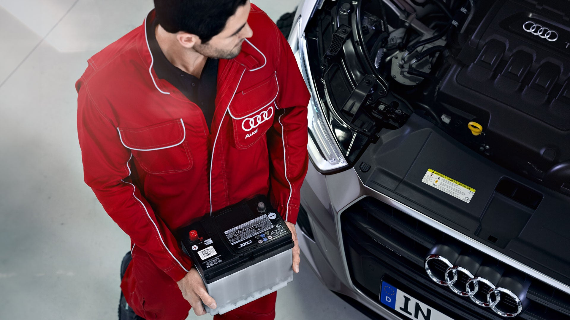 Audi-Mechaniker in roter Arbeitskleidung hält eine Autobatterie neben einem Fahrzeug mit geöffneter Motorhaube.   