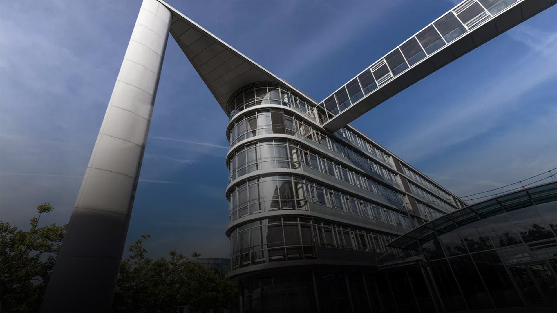 Modern office building with glass bridge under a blue sky, striking architecture with supporting column.