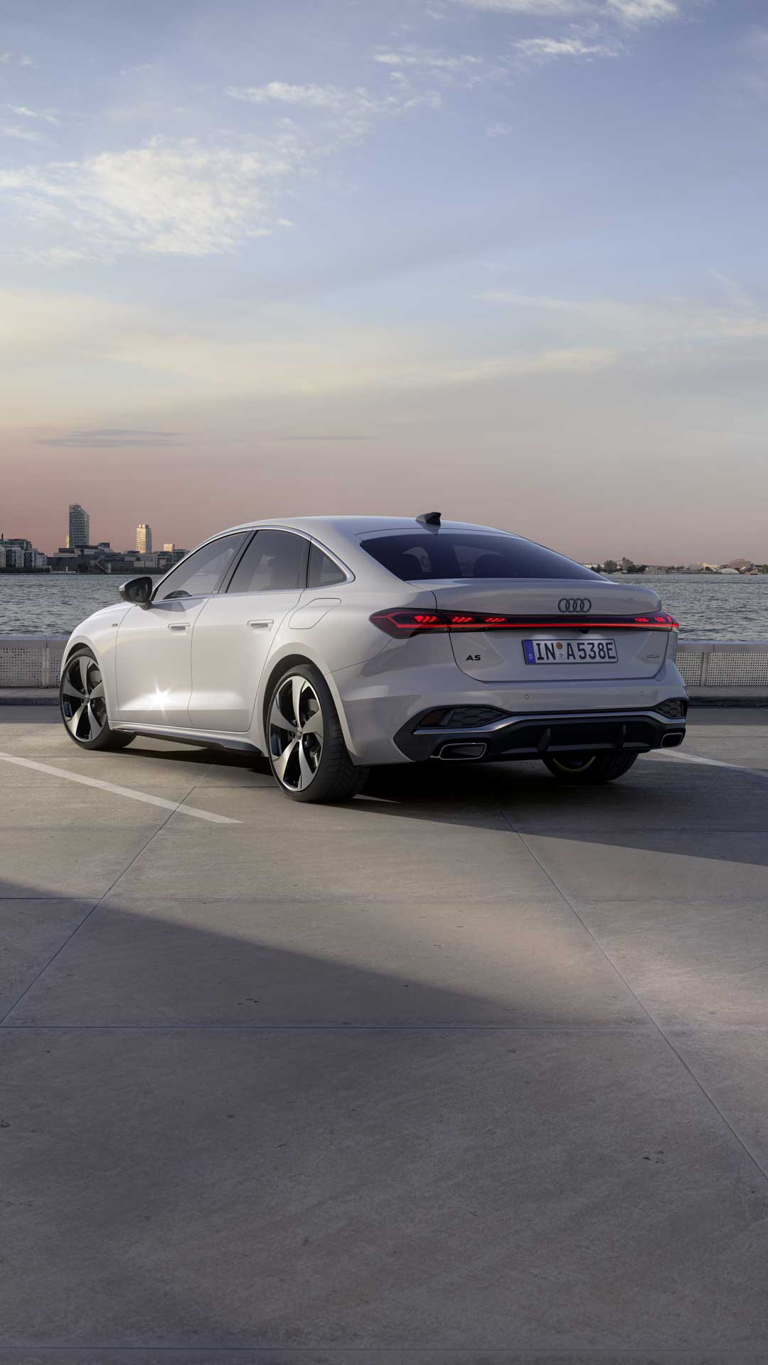 Die A5 Limousine e-hybrid steht im schrägen Heckprofil auf einem Parkplatz am Ufer eines Sees. Dahinter ist die Skyline einer Stadt bei Dämmerung zu sehen.