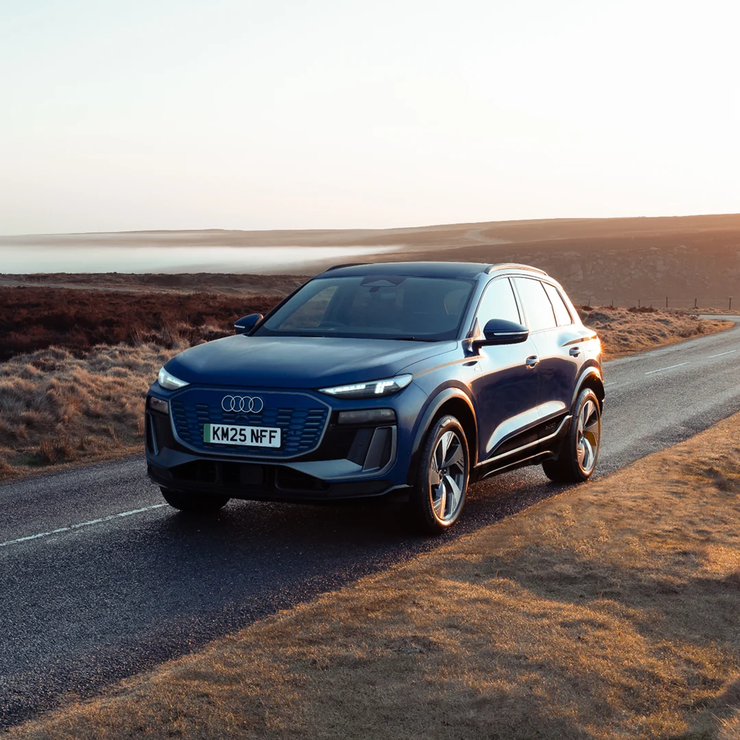 A dark blue Audi SUV drives along a coastal highway at sunset, with a scenic ocean and mountain backdrop. The scene conveys freedom and adventure.