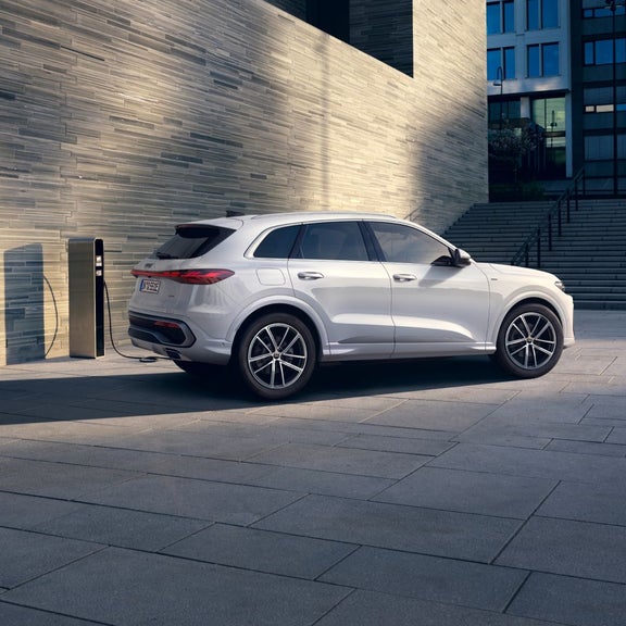 White SUV parked next to a charging station, set against a modern stone wall and urban architecture in the background.