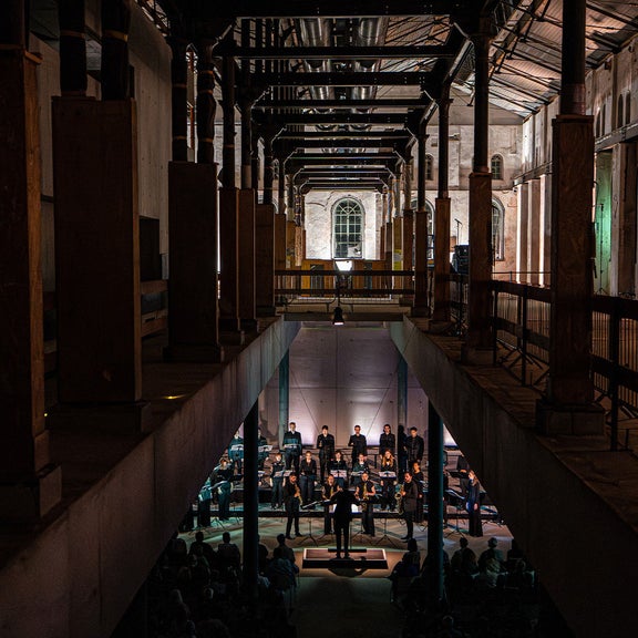 Chor und Dirigent in einer dramatisch beleuchteten Industriehalle mit hohen Säulen und Publikum.