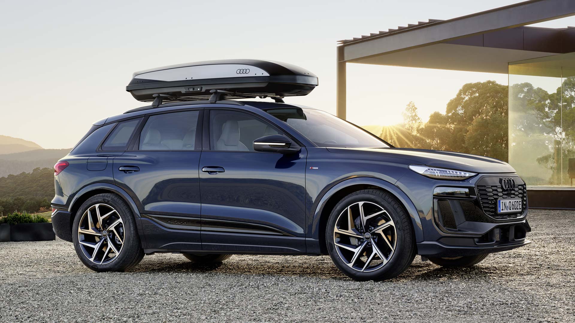 The Q6 SUV e-tron with roof box is parked sideways on the gravel area in front of a modern house, illuminated by the setting sun.