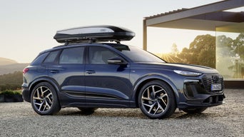 The Q6 SUV e-tron with roof box is parked on a gravel area in front of a modern house at sunset.