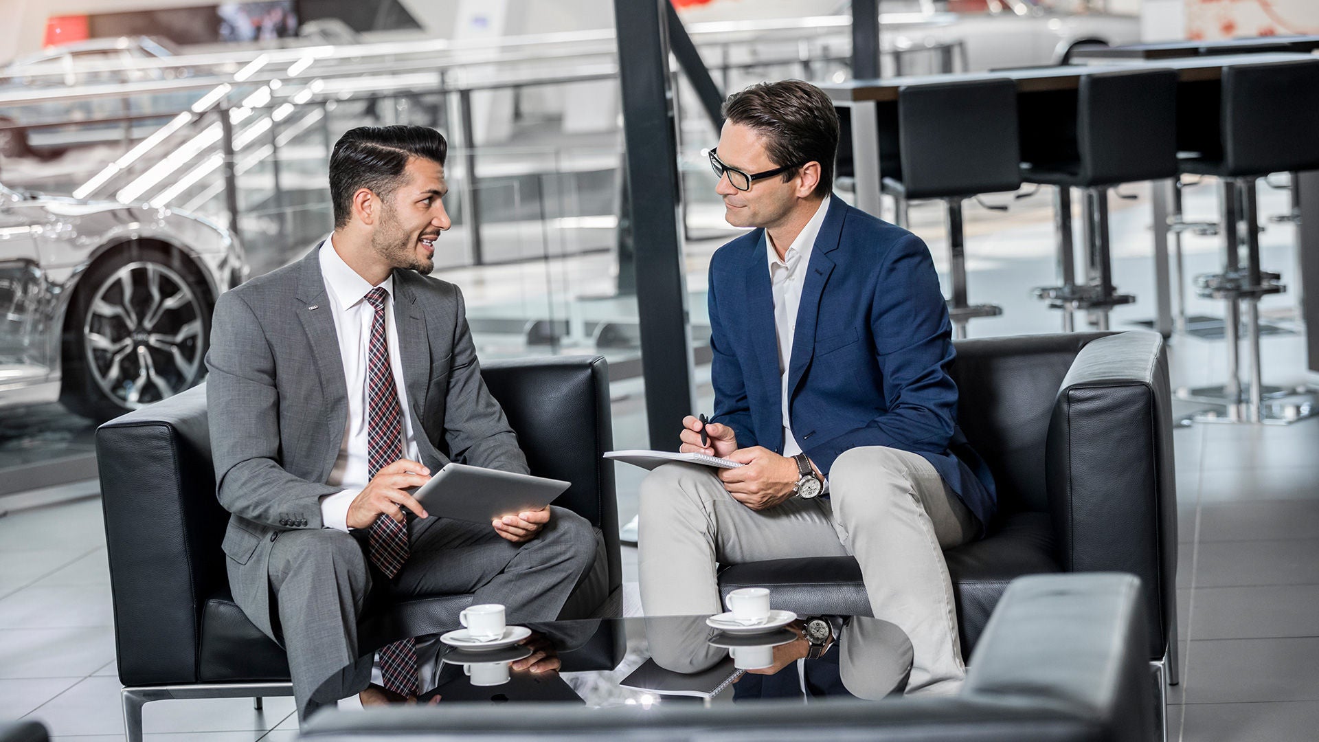 Zwei Männer in Anzügen sprechen mit Tablets und Kaffee in einem modernen Autohaus.