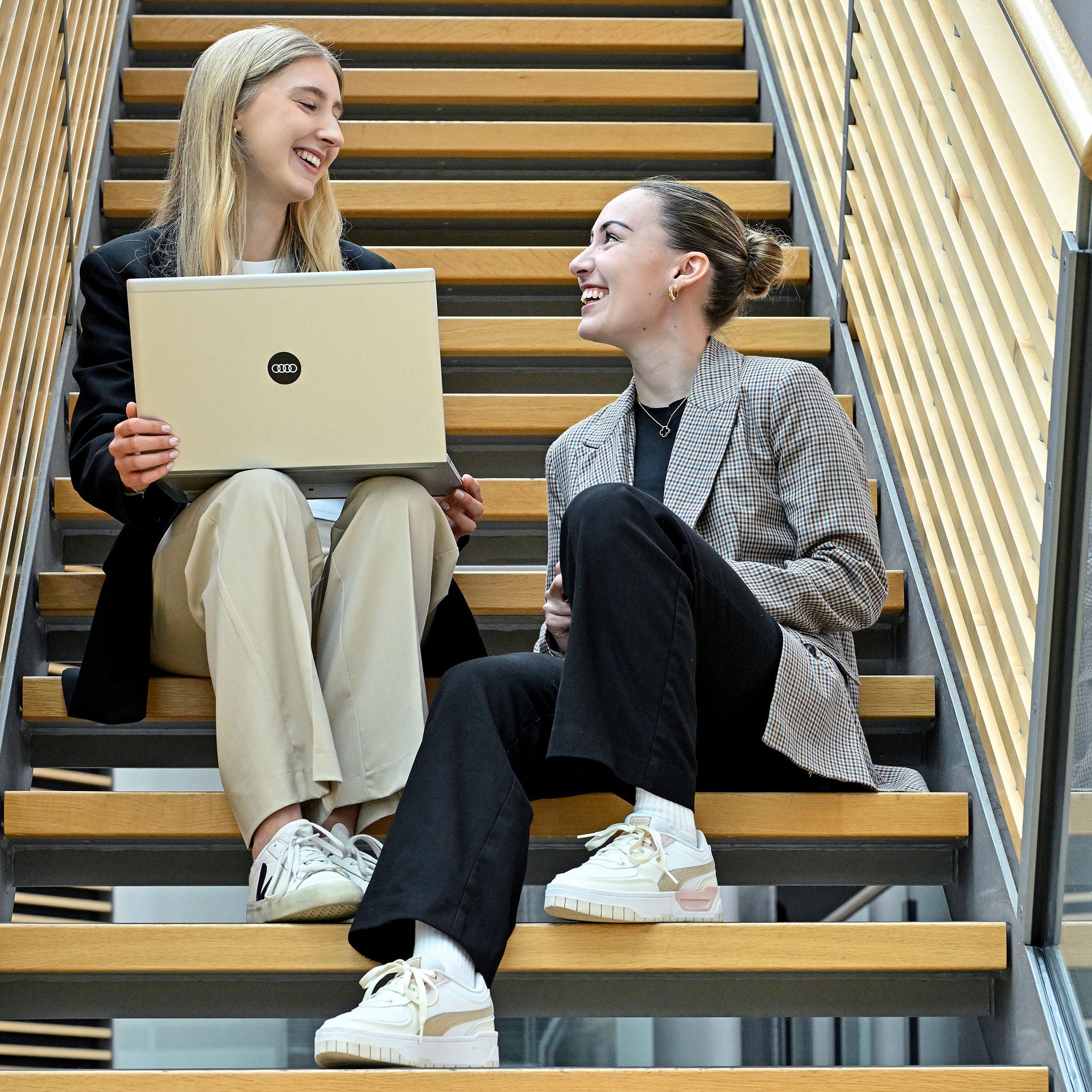 Zwei Mitarbeitende sitzen mit einem Laptop auf einer Treppe und lächeln sich an.