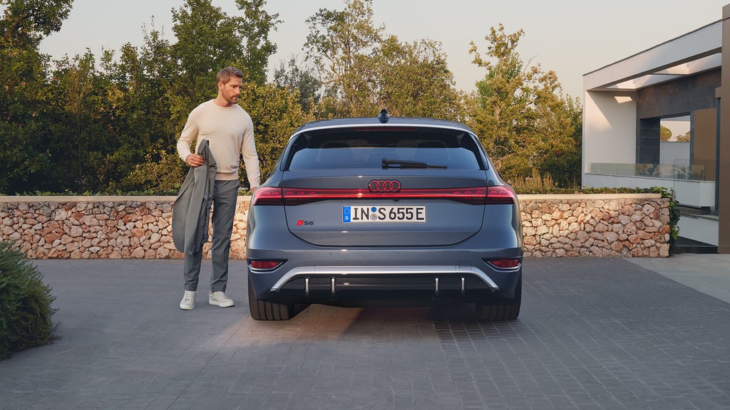 Ein Mann steht links neben einem dunkelblauen Audi S6 Avant e-tron, Rückansicht, auf einer gepflasterten Einfahrt vor einer Steinmauer.