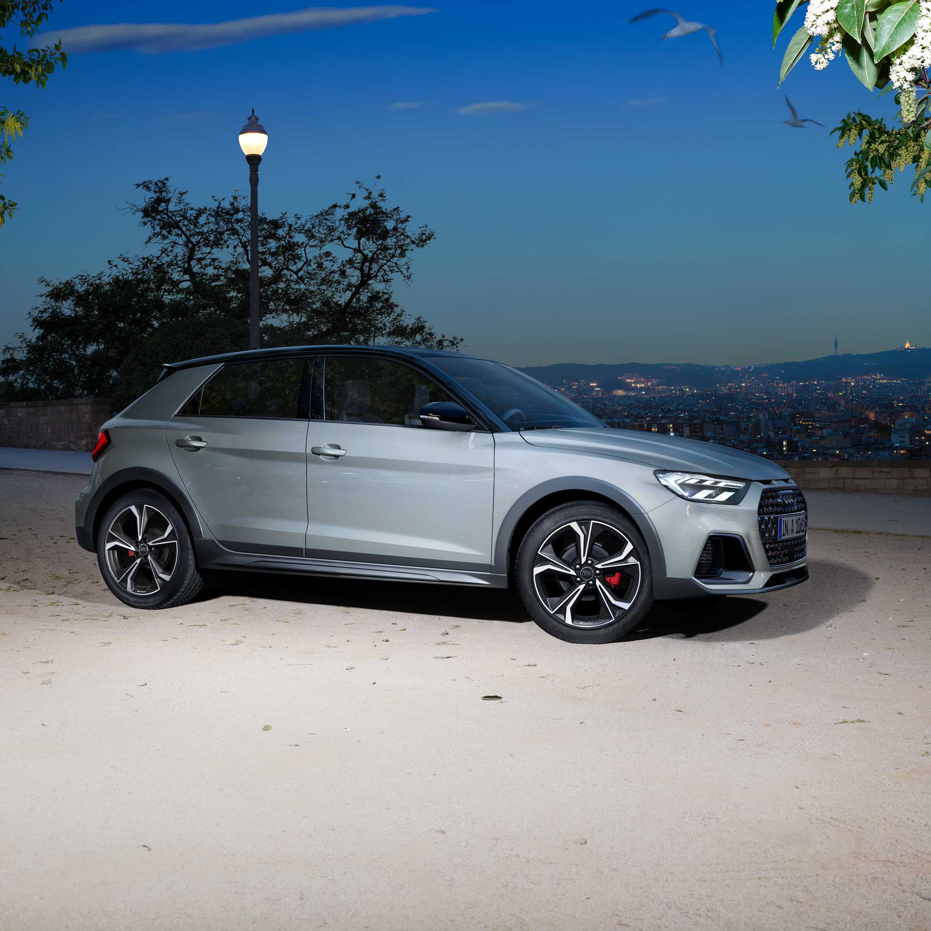 Silberner Audi A1 allstreet im Profil, geparkt auf heller Fläche mit Blick auf beleuchtete Stadt bei Nacht.