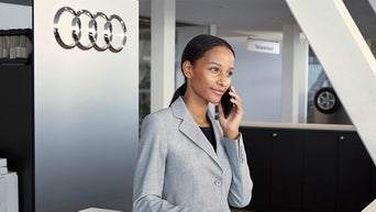 Frau im grauen Blazer telefoniert im Audi Autohaus, neben ihr ein Schild mit Audi Ringen.