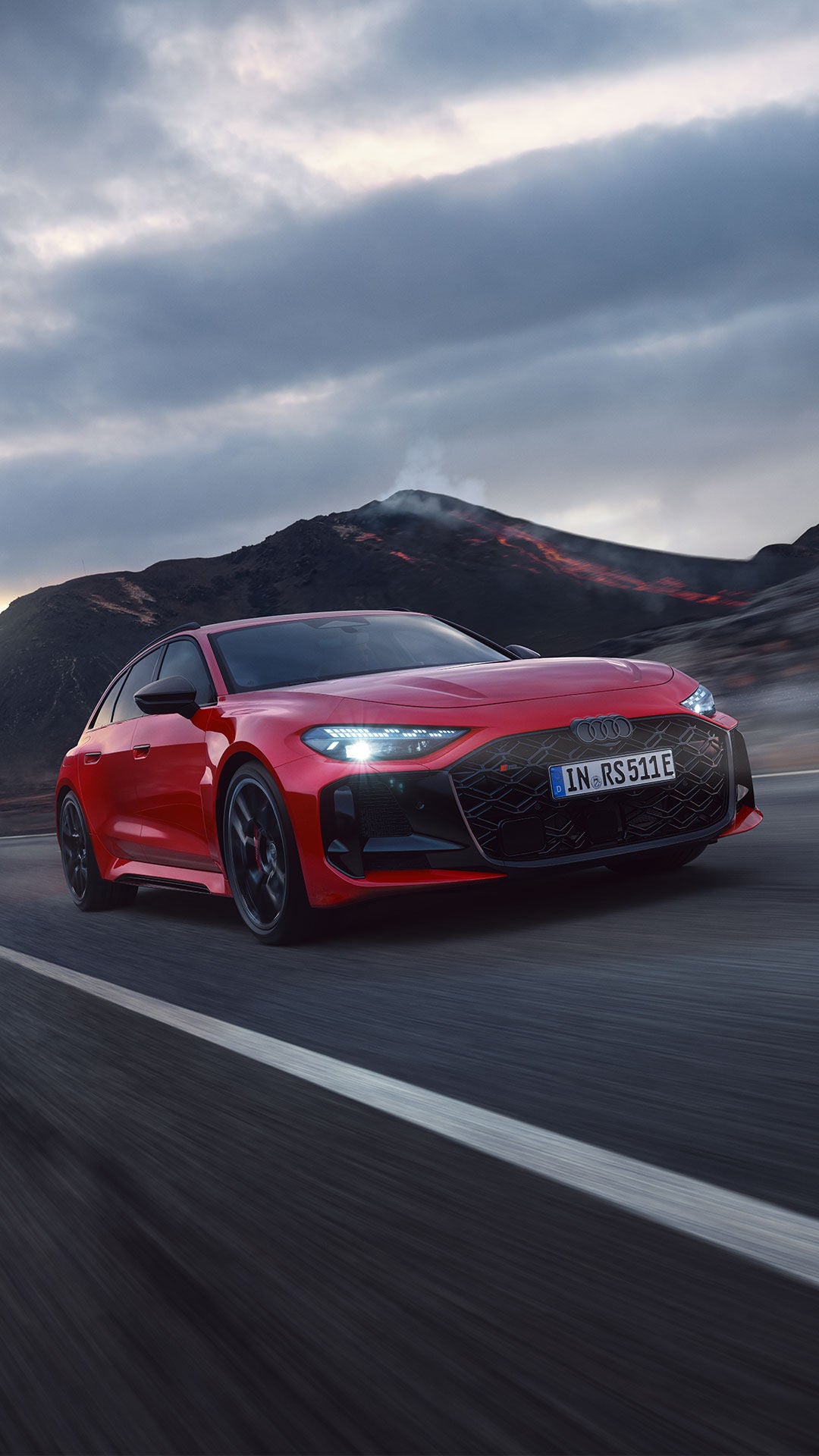 Roter Audi RS5 auf einer kurvenreichen Straße vor einer vulkanischen Berglandschaft. Im Hintergrund sind dunkle Lavahänge, Rauch und glühende Lava unter einem bewölkten Himmel zu sehen.