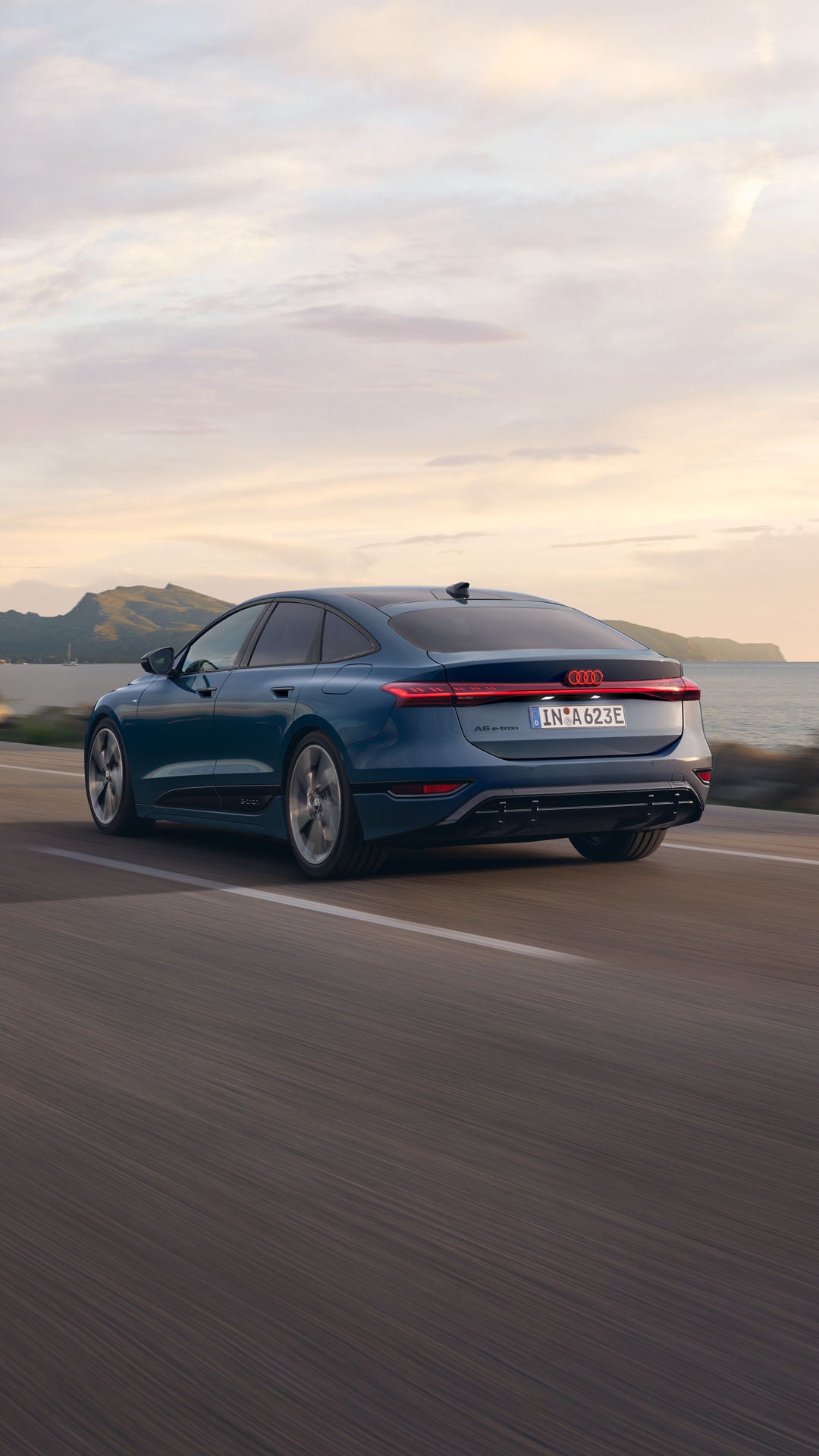 Der A6 Sportback e-tron aus schräger Heckansicht fährt eine Straße parallel zum Ufer entlang. Im Hintergrund sind Berge zu erkennen.