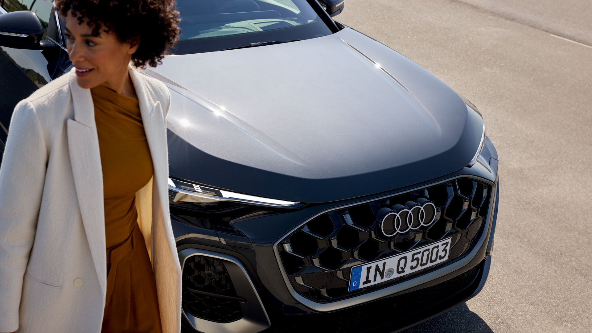 Frau in weißem Mantel steht lächelnd vor der Front eines schwarzen Audi Q5 mit Waben-Kühlergrill, aufgenommen bei Sonnenlicht.