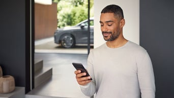 Mann in hellem Pullover steht im Haus und blickt lächelnd auf sein Smartphone.