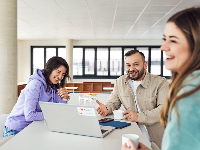 Buenyamin looks into the camera and sits at a table with colleagues. There is a laptop in the middle