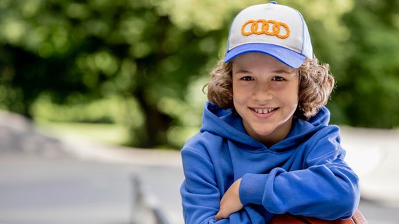 Lächelndes Kind mit blauem Hoodie und weiß-blauer Audi-Kappe vor grünem Hintergrund.