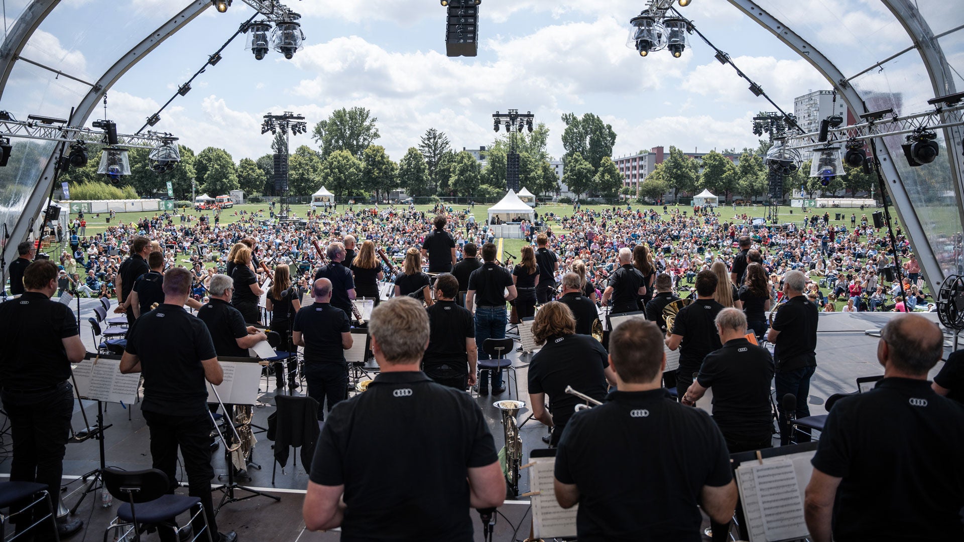 Orchester auf Open-Air-Bühne spielt vor großem Publikum im Park bei sonnigem Wetter.