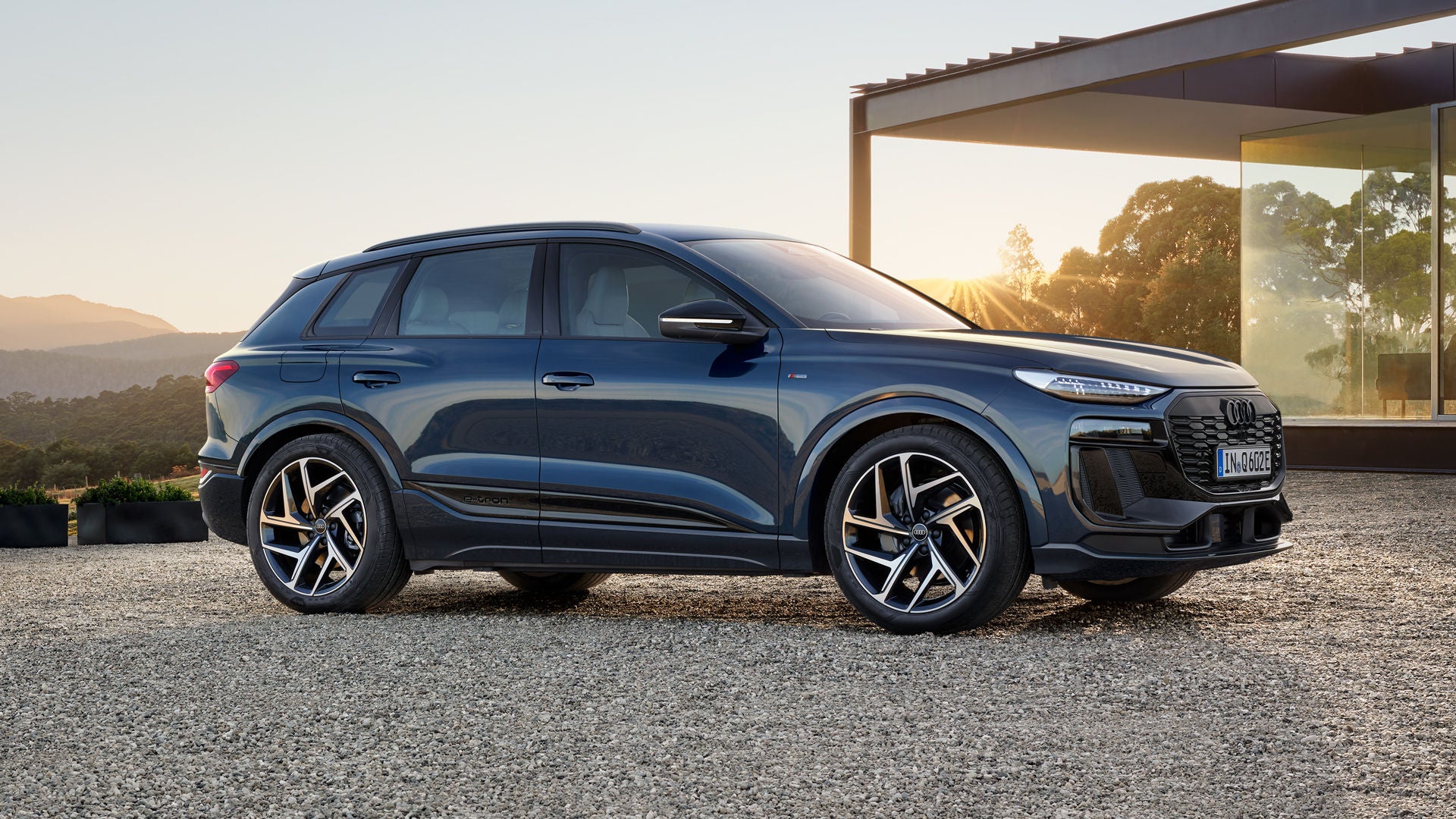 Der Q6 SUV e-tron steht seitlich auf der Kiesfläche vor einem modernen Haus und wird dabei von der untergehenden Sonne angestrahlt.