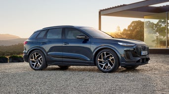Der Q6 SUV e-tron parkend auf steinigem Untergrund, dahinter ein rundum verglastes Gebäude. Die untergehende Sonne beleuchtet die Szenerie.