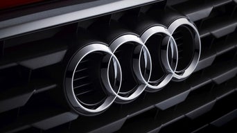 Nahaufnahme des Audi-Logos mit vier Ringen auf schwarzem Kühlergrill eines Fahrzeugs.