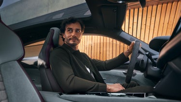 A young man with a three-day beard and dark curls sits in an Audi model and looks to the side. In the background is a wooden panel wall