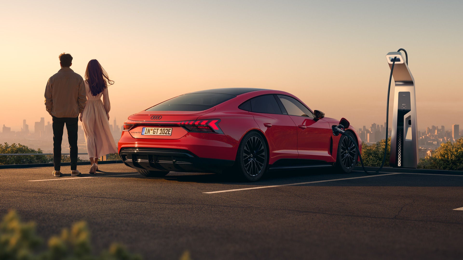 Roter Audi e-tron GT quattro lädt an einer Ladesäule bei Sonnenuntergang. Neben dem Fahrzeug stehen ein Mann und eine Frau mit dem Rücken zur Kamera gerichtet.