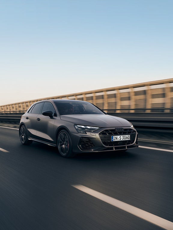 Grauer Audi S3 Sportback fährt auf leerer Straße an Betonbarriere entlang, Blick auf Front und linke Seite.