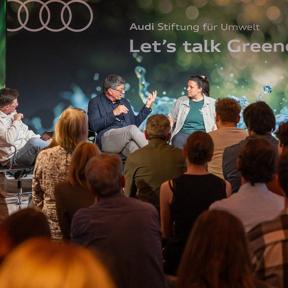 Four people discussing on a stage with an audience in the foreground. A picture of a monkey can be seen on the left of the screen.