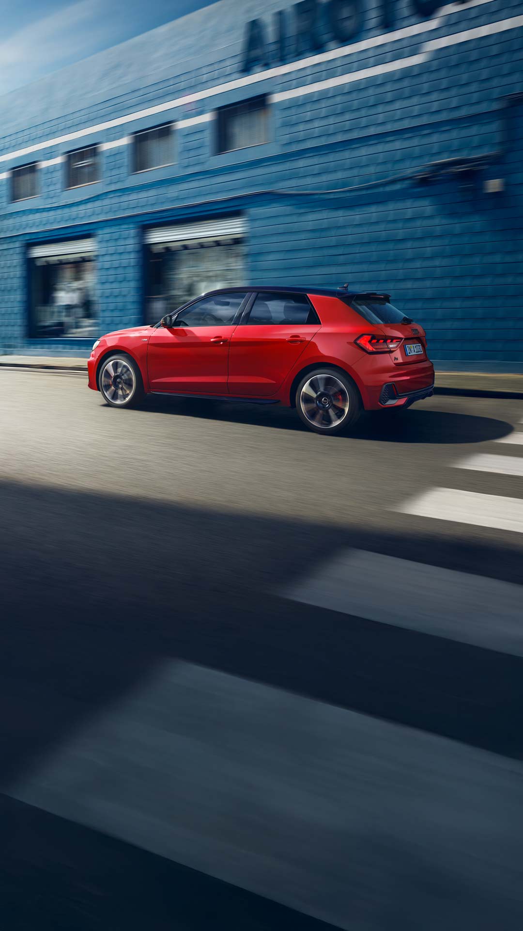 Roter Audi A1 Sportback fährt auf einer Straße vor einer blauen Wand, Seitenansicht von links.