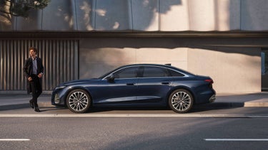 Ein Mann im dunklen Mantel geht auf dem Gehweg an einer blauen Audi A6 Limousine vorbei, die vor einer Betonwand mit vertikalen Lamellen geparkt ist.