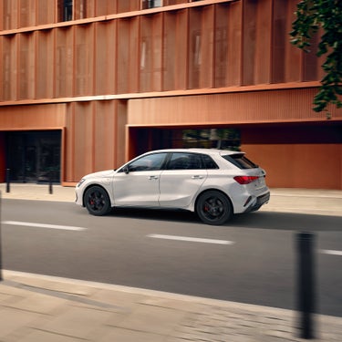 Weißer Audi A3 Sportback TFSI e fährt auf städtischer Straße an moderner, orangefarbener Gebäudefassade vorbei.