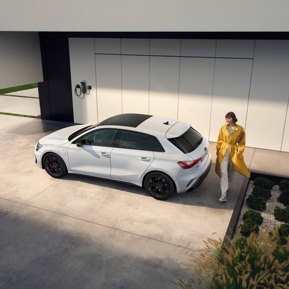 A woman in a yellow coat stands next to a white car parked on a driveway. The setting is modern and clean, with a sleek wall-mounted charger visible.