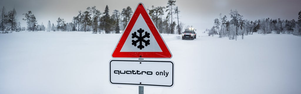 Schneeflächenszene mit Verkehrsschild „quattro only“ und Auto im Hintergrund.
