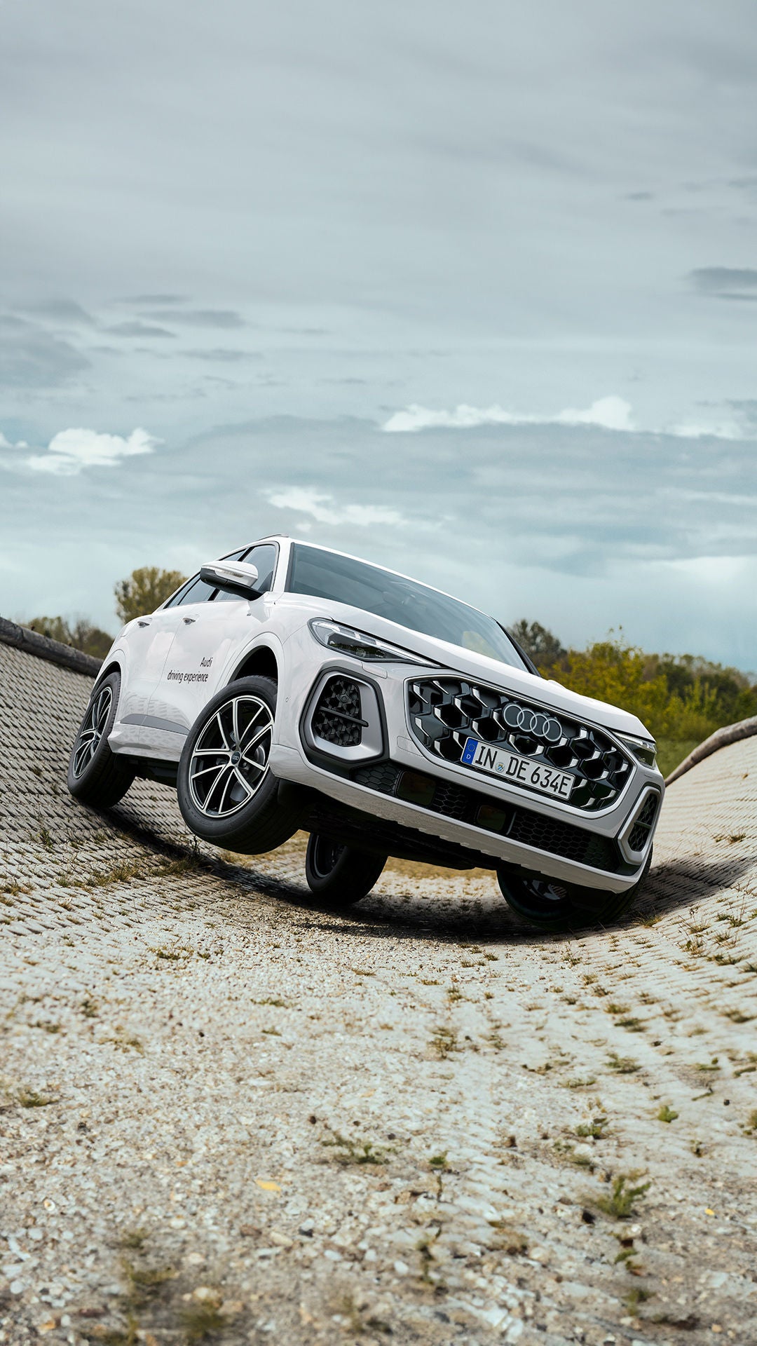 Weißer Audi Q5 SUV e-hybrid in geneigter Kurve auf gepflasterter Teststrecke; Front/Seite sichtbar, bewölkter Himmel.