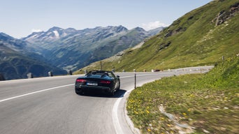 Ein schwarzer Audi R8 Spyder V10 performance quattro mit offenem Verdeck (Heckansicht) fährt durch eine scharfe Kurve in alpiner Landschaft bei klarem Himmel.