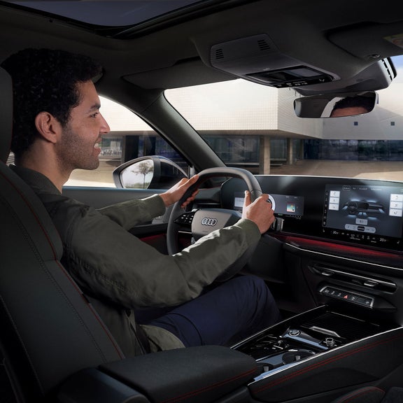 A person driving an Audi Q3, showing the modern dashboard, steering wheel, and touchscreen controls.