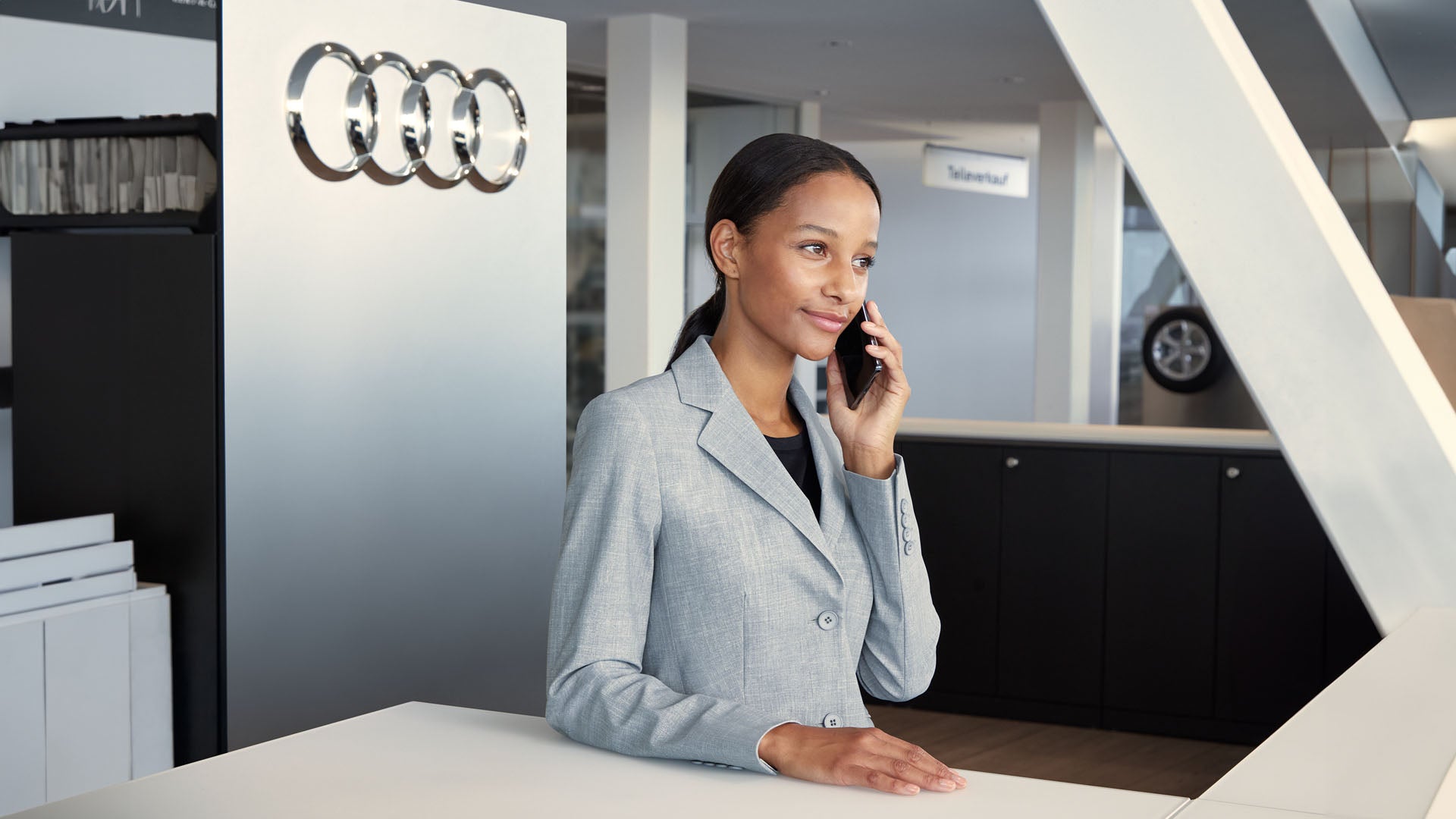 Frau im grauen Blazer telefoniert an einem Empfangstresen mit Audi-Logo im Hintergrund.