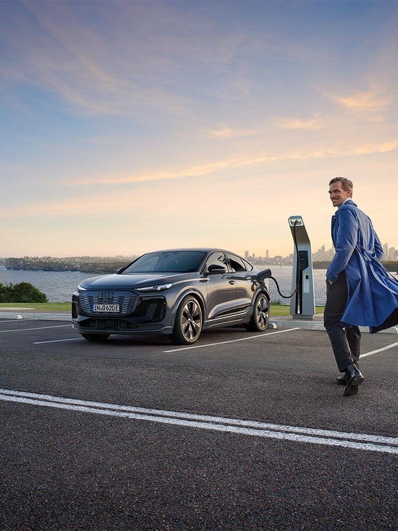 Ein Mann in blauem Mantel läuft zu einem ladenden Audi Q6 Sportback e-tron und lächelt zur Seite, im Hintergrund Skyline und Wasser.