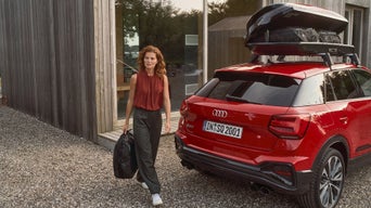 Frau mit Tasche geht neben rotem Audi mit geöffneter Dachbox vor Holzhaus.