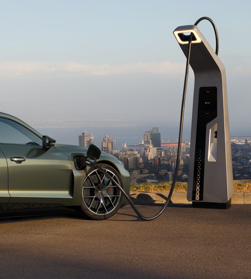 Um Audi RS e-tron GT performance verde está carregando em uma estação de carregamento rápido com vista para a cidade ao fundo.