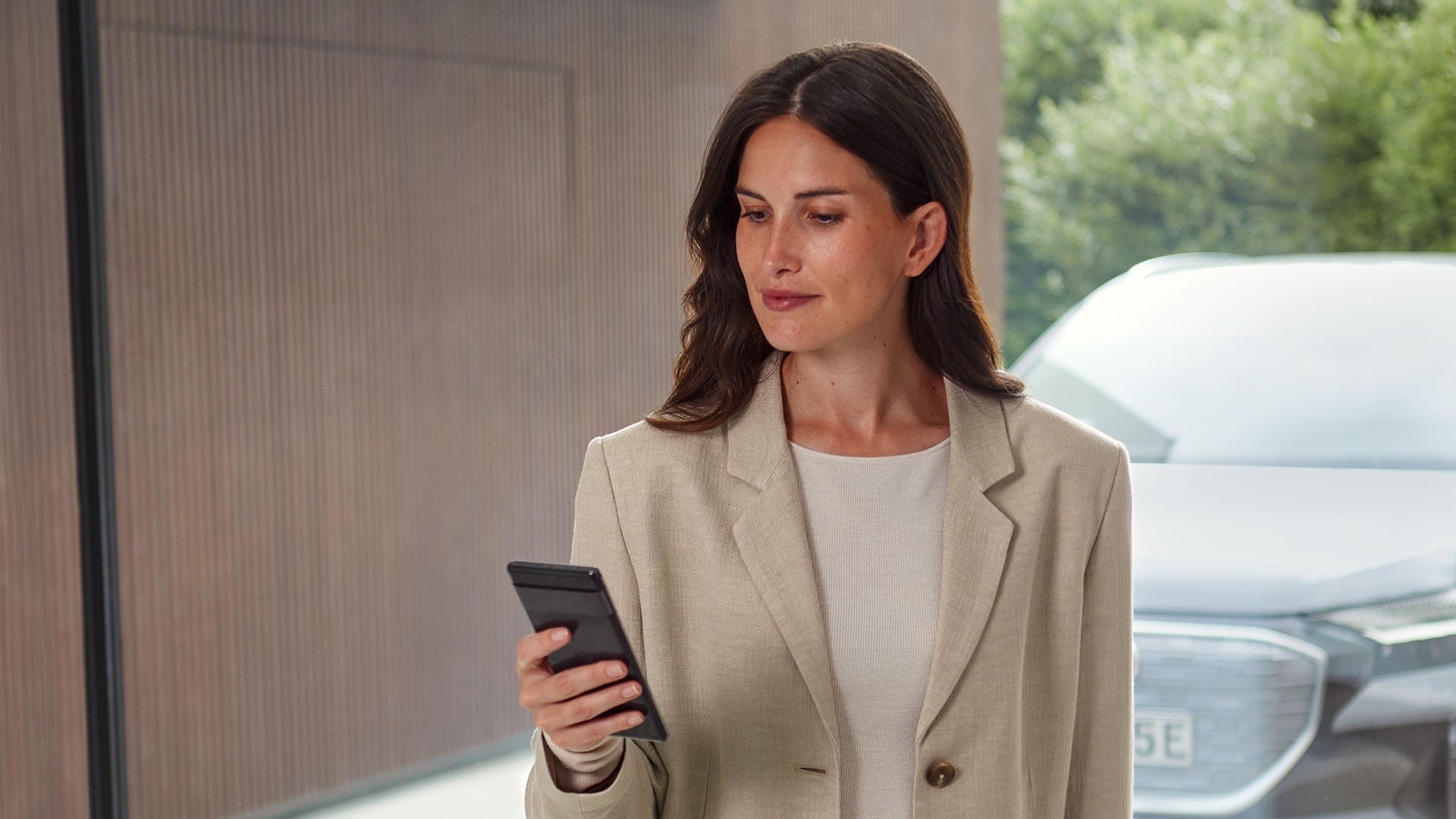 Eine Frau im beigen Blazer blickt auf ihr Smartphone, im Hintergrund steht ein silberner Audi.